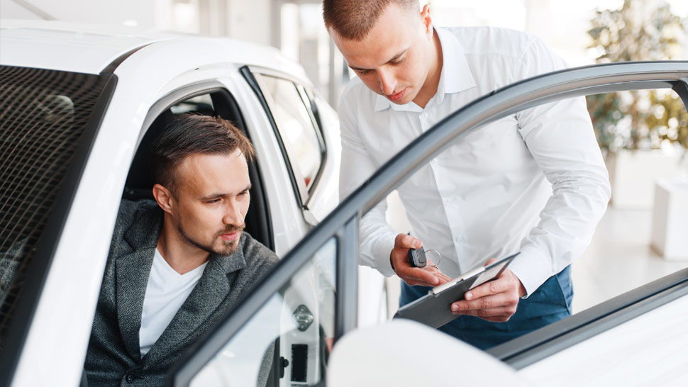 Na sliki sta dva moška ob belem avtomobilu. Eden sedi na voznikovem sedežu, drugi pa stoji zunaj in mu na tablici razlaga ali prikazuje dokument. Oba sta osredotočena na pogovor.