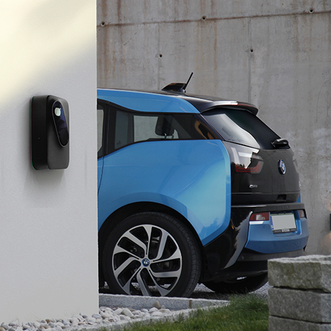 Na sliki je modro-črn električni avtomobil, parkiran ob hiši in priključen na stensko domačo polnilnico. Polnilnica je nameščena na svetli fasadi, v ozadju pa je betonski zid in okolica z zelenico in kamnitimi elementi.