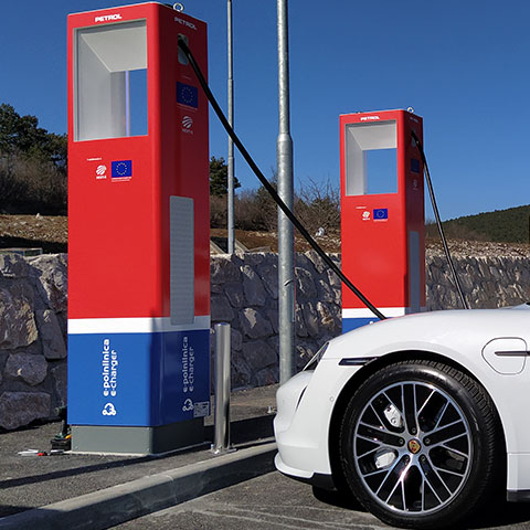 Na sliki je belo električno vozilo, priključeno na eno od dveh Petrolovih javnih polnilnic ob kamnitem zidu, z značilnim rdeče-modrim ohišjem. Na polnilnicah so vidni logotipi Evropske unije in oznake projekta e-polnilnice.