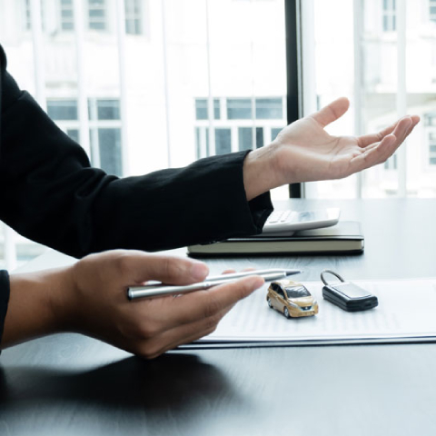 Na sliki sta dve osebi v poslovnem okolju. Ena oseba sedi za mizo in drži pisalo, medtem ko druga z gesto rok pojasnjuje ali razlaga nekaj v zvezi z dokumenti na mizi. Na dokumentih sta razstavljena majhen model avtomobila in avtomobilski ključ.