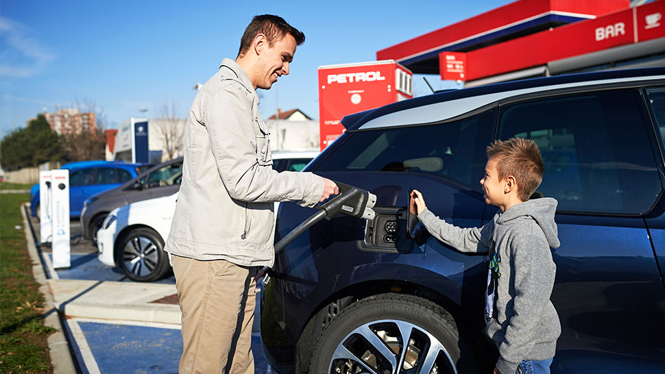 Na sliki moški polni električni avtomobil na Petrolovi polnilnici, ob njem pa stoji deček, ki ga z zanimanjem opazuje. Oba se nasmihata, kar ustvarja prijetno in sproščeno vzdušje. V ozadju so vidne še druge polnilnice in Petrolov bencinski servis.