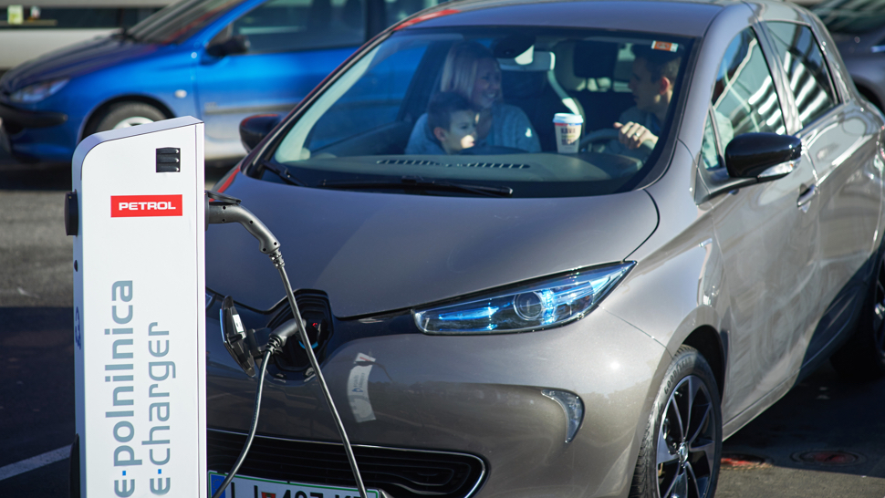 Na sliki je električni avtomobil, priključen na Petrolovo polnilnico z oznako »e-polnilnica«. V vozilu sedi družina – odrasla oseba in otrok – ki se med polnjenjem sproščeno pogovarjajo. V ozadju so parkirani avtomobili.