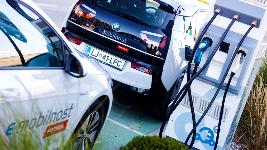 Na sliki sta dve električni vozili z oznako »e-mobilnost« in logotipom Petrol, parkirani ob polnilni postaji. Vozili se polnita na beli električni polnilnici z več priključki in kabli. V ospredju je vidna modro-bela polnilna postaja.