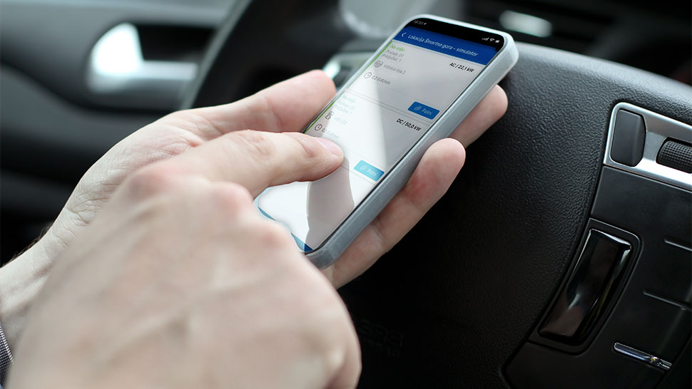 Na sliki je oseba, ki v avtomobilu uporablja pametni telefon z odprto aplikacijo OneCharge. Na zaslonu so prikazane informacije o prostih polnilnih mestih in moči polnjenja. V ozadju je volan vozila, kar poudarja enostaven.