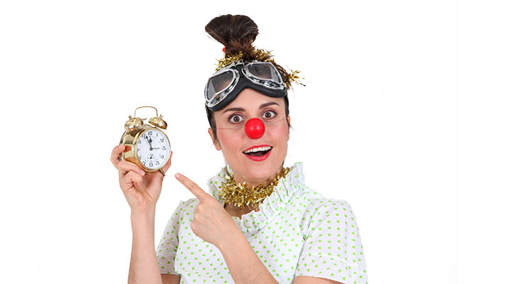 Klovnesa z rdečim nosom in zaščitnimi očali na glavi pred belim ozadjem drži starinski budilnik in z drugo roko kaže nanj.