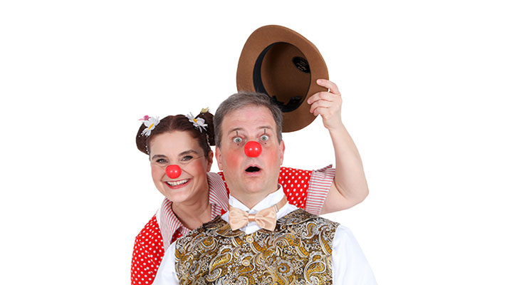 Dva klovna z rdečimi noski pred belim ozadjem; spredaj stoji presenečen moški, zadaj nasmejana ženska dviguje njegov klobuk.