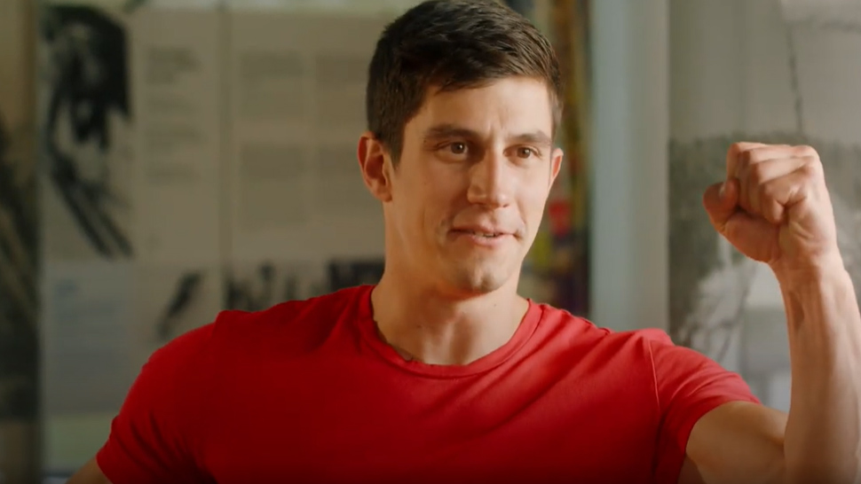 Moški v rdeči majici z nasmeškom dvigne obe stisnjeni pesti v zmagovalni pozi.