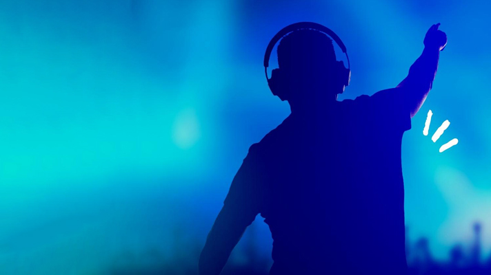 DJ s slušalkami dvignjeno roko vodi množico v klubu, osvetljen z modro svetlobo in dinamičnim vzdušjem.