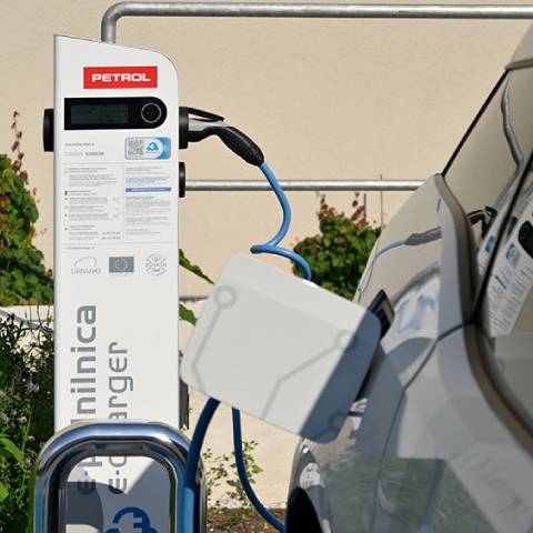 Na sliki je električna polnilnica Petrol z belim ohišjem in priklopljenim modrim kablom, ki polni električno vozilo. Na polnilnici so vidni logotipi Petrol, Evropske unije in programa URBANE.