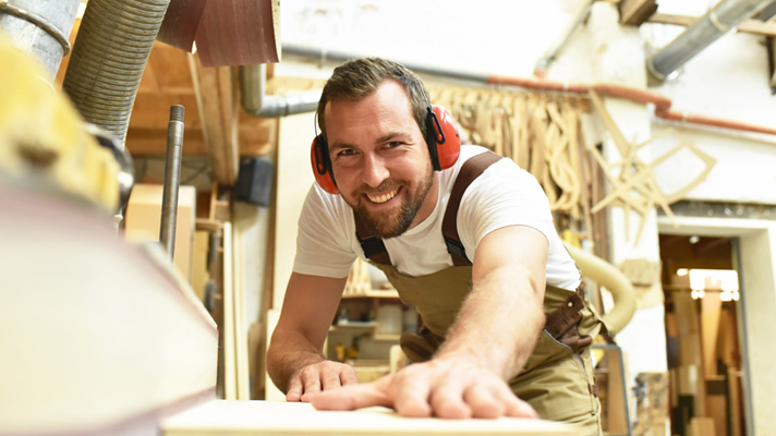Nasmejan moški v delovnem kombinezonu z zaščitnimi ušesnimi slušalkami dela v lesarski delavnici, obdano z lesenimi izdelki in orodjem.