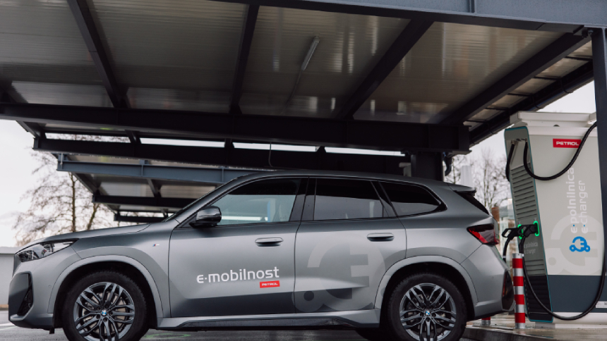 Sivo električno vozilo z oznako »e-mobilnost« in logotipom Petrol je priključeno na hitro polnilnico pod nadstreškom. Na polnilnici je vidna Petrolova oznaka »e-polnilnice charger«. Vozilo se polni na urejenem parkirnem prostoru, v ozadju pa se vidi del zunanjega okolja z drevesi.