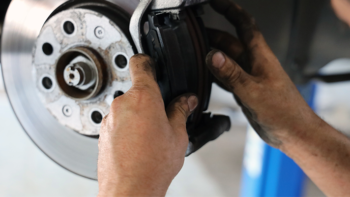 Na sliki so roke mehanika, ki namešča ali popravlja zavorne ploščice na avtomobilskem kolesu. Vidni so zavorni disk in del kolesne osi.