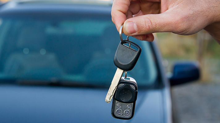 Na sliki je roka, ki drži avtomobilski ključ z daljincem. V ozadju je zamegljen avtomobil.
