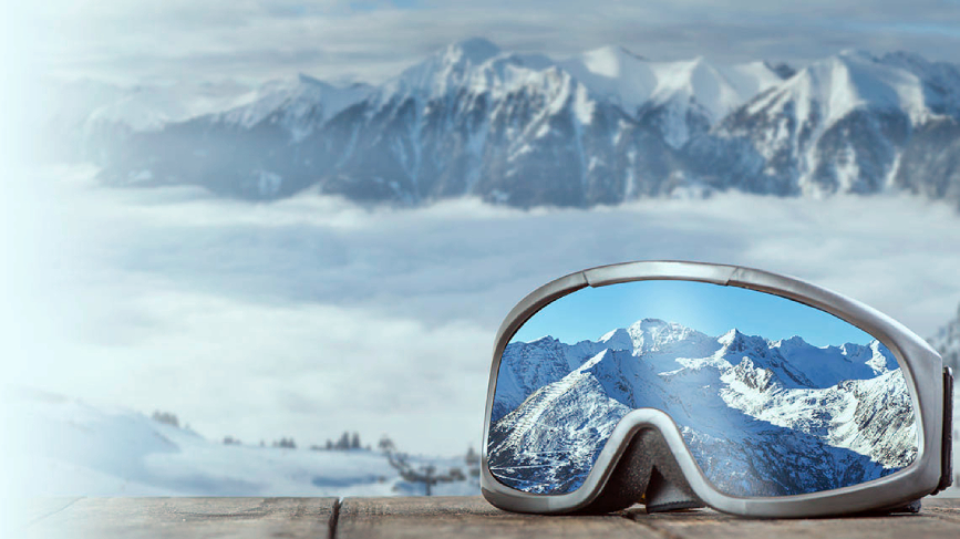Na sliki so smučarska očala na leseni podlagi. V steklu očal se odseva jasen pogled na zasnežene gorske vrhove, medtem ko je v ozadju enako prikazano zasneženo gorovje obdano z oblaki in meglo.