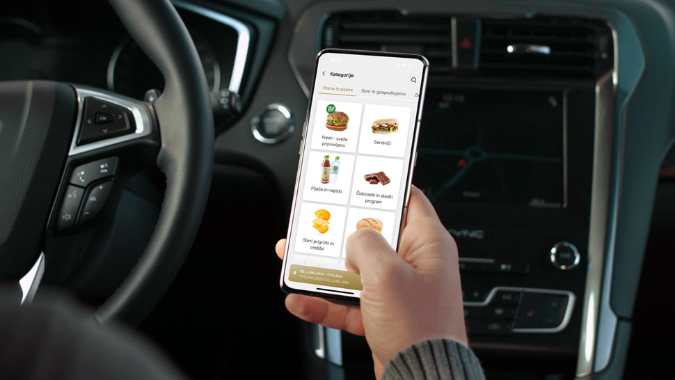 Oseba v avtomobilu drži pametni telefon z odprto aplikacijo Petrol GO, kjer so prikazane kategorije izdelkov, kot so sendviči, pijače, sladice in prigrizki.