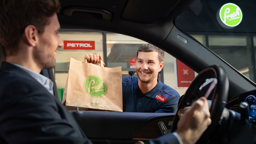 Prodajalec prodajnega mesta Petrol prinese papirnato vrečko z napisom Fresh stranki, ki sedi v avtomobilu in prevzema naročilo na Petrolu.