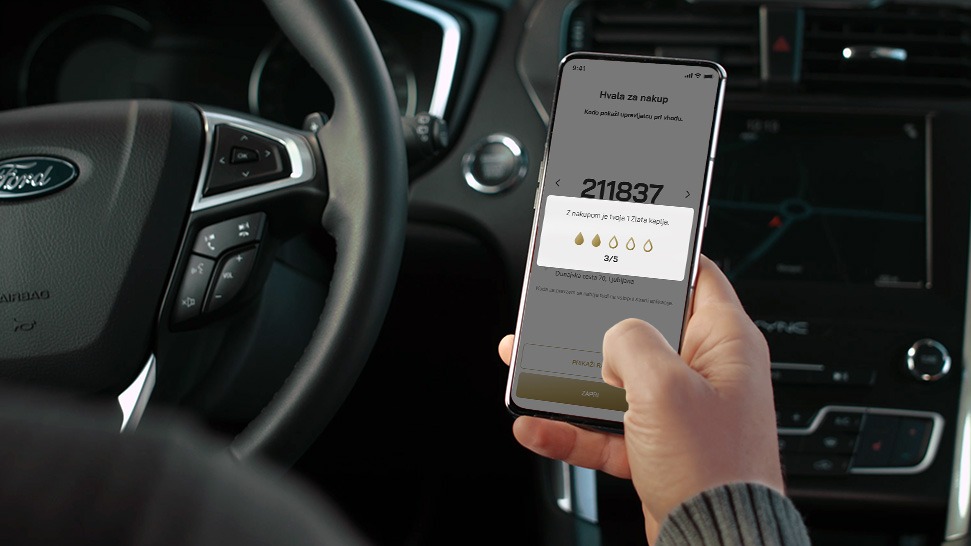 Na sliki je voznik v avtomobilu, ki v roki drži pametni telefon z odprto aplikacijo Petrol. Na zaslonu je prikazana številka računa in možnost pregleda zbranih kapelj od avtopranj.