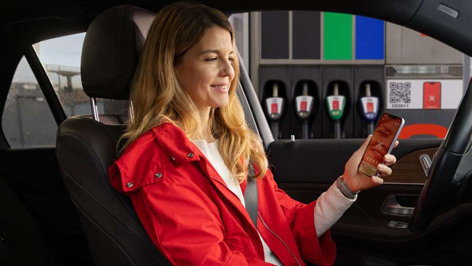 Ženska v rdeči jakni sedi v avtomobilu pred bencinsko črpalko in na telefonu uporablja Petrol aplikacijo z odprto QR kodo, v ozadju pa so vidne ročke za točenje goriva.