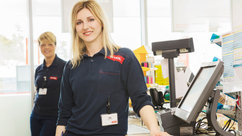 Na sliki sta dve Petrolovi prodajalki v temno modri uniformi z rdečim logotipom Petrol. Ena izmed njiju stoji ob blagajni in se nasmejano obrača proti kameri, druga pa stoji v ozadju in prav tako prijazno gleda naprej.