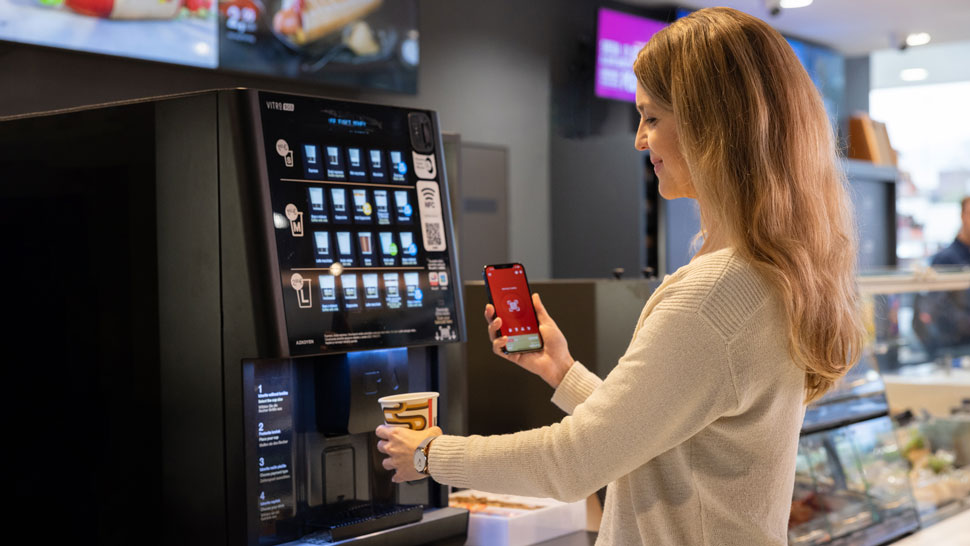 Ženska si pripravlja kavo na kavomatu, medtem ko uporablja Petrol aplikacijo na telefonu za skeniranje in plačilo z QR kodo.