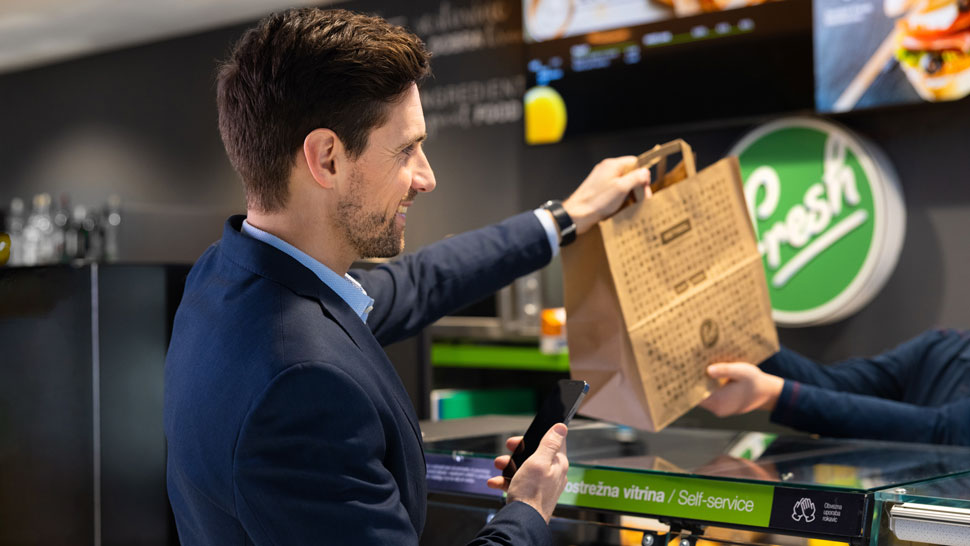 Na sliki je moški v poslovni obleki, ki v Petrol Fresh prodajalni prevzema papirnato vrečko z naročenimi izdelki. V eni roki drži pametni telefon, v drugi pa sprejema vrečko, ki mu jo izroča prodajalec. V ozadju je viden logotip Fresh.