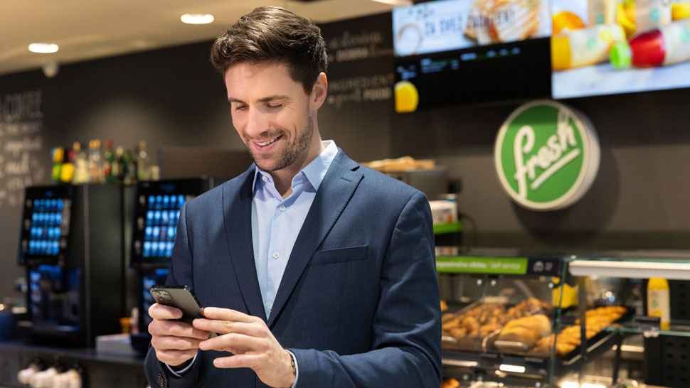 Moški v poslovni obleki uporablja pametni telefon v Petrol prodajalni, kjer so v ozadju vidni prigrizki, napitki in prepoznaven Fresh logotip