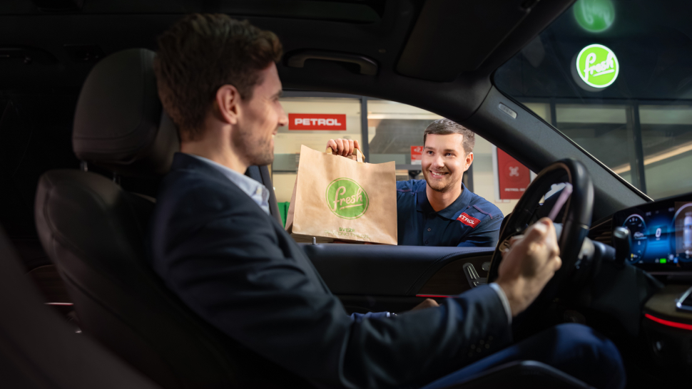 Na sliki prodajalec v Petrolovi uniformi skozi okno avtomobila izroča papirnato vrečko z logotipom Fresh nasmejanemu vozniku v poslovni obleki. V ozadju sta vidna logotipa Petrol in Fresh na stavbi.