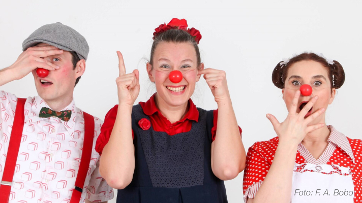 Trije nasmejani klovni z rdečimi noski pozirajo pred belim ozadjem in se igrivo dotikajo svojih nosov.