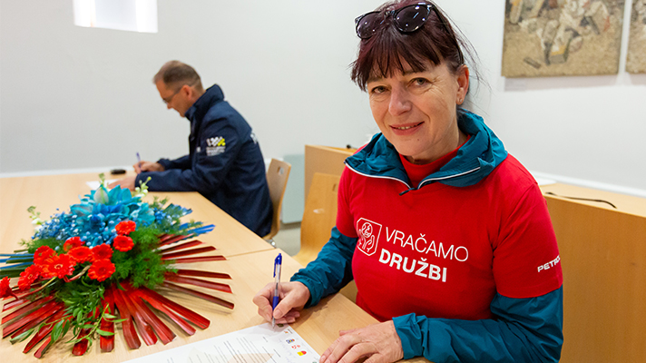 Ženska v rdeči majici »Vračamo družbi« za mizo podpisuje dokument, ob njej je šopek modro-rdečega cvetja, v ozadju sedi še en udeleženec.