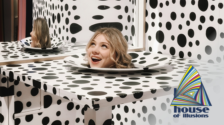 Na sliki je glava nasmejanega dekleta, ki je postavljena na krožniku mize s črno-belimi pikami, kar je del scene v Hiši iluzij. Desno spodaj se nahaja logotip 'House of Illusions'.