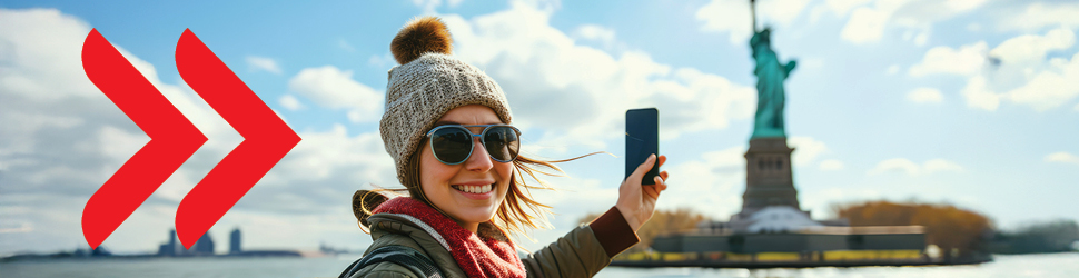 Na sliki je prikazana nasmejana oseba, ki na prostem ob vetrovnem vremenu s telefonom v roki dela »selfie« s kipom Svobode v ozadju. Oseba nosi zimsko kapo, sončna očala in šal, ob strani slike pa sta vidni dve veliki rdeči puščici, usmerjeni v desno. V ozadju je vodna gladina ter del mestne panorame pod svetlim, delno oblačnim nebom.
