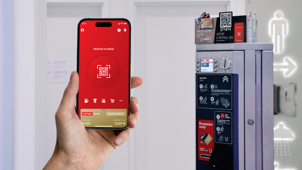 Fotografija prikazuje roko osebe, ki drži pametni telefon s prikazano aplikacijo Petrol. Na zaslonu je prikazana rdeča stran z napisom »Skeniraj in plačaj« ter QR kodo na sredini. V ozadju je kovinski avtomat za vstop v stranišče, ki ima pritrjene nalepke z informacijami in QR kodo za dostop. Na desni strani slike so vidne svetlobne oznake za smer in simbol osebe, ki označuje stranišče.