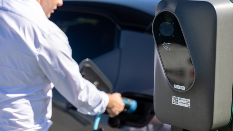 Na sliki je oseba, ki priklaplja električno vozilo na polnilnico podjetja Petrol. Polnilnica je v ospredju slike, medtem ko sta oseba in vozilo rahlo zamegljena v ozadju.