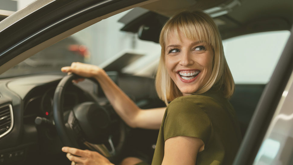 Nasmejana dekle s svetlimi lasmi sedi za volanom avtomobila, drži volan in se veselo ozira nazaj.