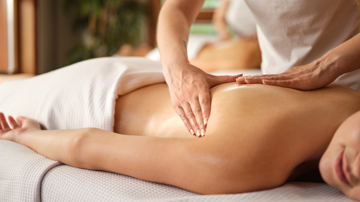 Slika prikazuje masažo hrbta, kjer oseba, ki leži na trebuhu, pokrita z belo brisačo, prejema masažo od rok terapevta. Oseba, ki prejema masažo, deluje sproščeno in se smehlja. Ozadje slike je zamegljeno in prikazuje notranjost spa centra.