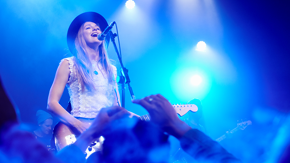 Pevka s kitaro na odru, oblečena v bel top in črn klobuk, poje v mikrofon med nastopom. Na odru jo obdaja modra svetloba, v ospredju pa so vidne dvignjene roke navdušene publike.