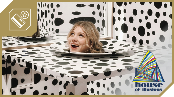 Na sliki je glava nasmejanega dekleta, ki je postavljena na krožniku mize s črno-belimi pikami, kar je del scene v Hiši iluzij. Rob slike je obarvan zlato, zgoraj levo se nahaja ikona kartice z zvezdico, desno spodaj pa logotip 'House of Illusions'.