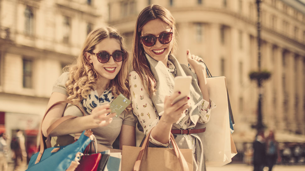 Na sliki sta dve nasmejani mladi ženski na prostem, ki uživata v nakupovanju. Obe nosita sončna očala in držita nakupovalne vrečke. Ženska na levi v eni roki drži novo Petrol Pay Loyalty plačilno kartico, v drugi pa torbico, medtem ko se nagiba proti prijateljici. Ženska na desni drži pametni telefon in z njim nekaj prikazuje ali fotografira. V ozadju je mehko zamegljeno mestno okolje z večjo zgradbo. Slika ponazarja brezskrbno nakupovanje in uporabo Petrol Pay Loyalty kartice tudi izven Petrolovih prodajnih mest.