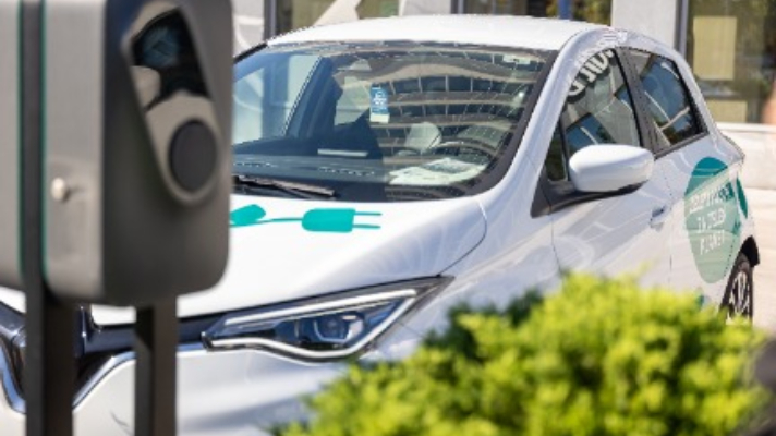 Na sliki je belo električno vozilo z zelenimi oznakami v obliki električnega vtiča, parkirano ob polnilnici. V ospredju je del naprave za polnjenje, obdan z zelenjem.