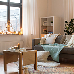 Na sliki je svetla dnevna soba z okrasnimi lučkami na okenski polici, sivim kavčem z odejami in blazinami ter majhno leseno klubsko mizico.