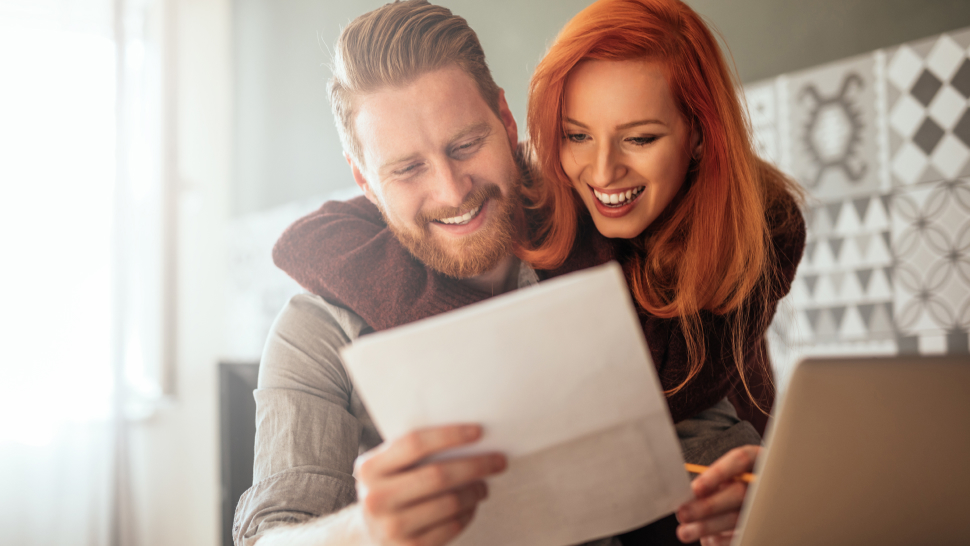 Na sliki sta nasmejana moški in ženska, ki skupaj gledata dokument. Sedita za mizo z odprtim prenosnikom, videti pa je, da sta zadovoljna z vsebino papirja, ki ga držita.