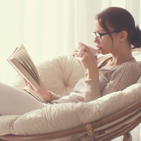 Na sliki je ženska z očali, ki sedi v naslanjaču, bere knjigo in pije iz skodelice.