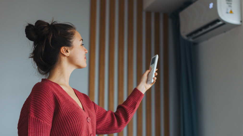 Ženska v rdečem puloverju z daljinskim upravljalnikom v roki upravlja klimatsko napravo, nameščeno na steni dnevnega prostora.