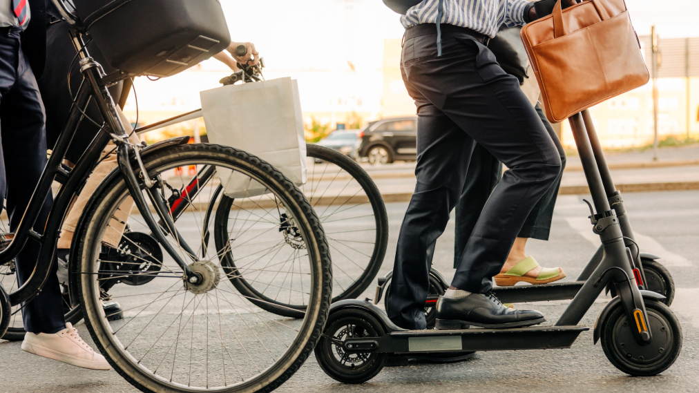 Na sliki so ljudje v poslovnih oblačilih, ki se peljejo z električnimi skiroji in kolesi po mestni ulici.