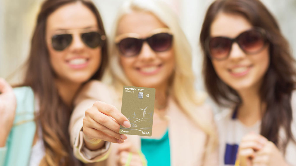 Tri nasmejane dekleta z sončnimi očali, ena izmed njih v ospredju drži zlato kartico Petrol Pay Loyalty Visa.