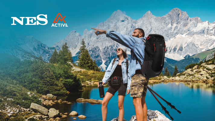 Dve osebi stojita ob alpskem jezeru s pohodniško opremo; ena oseba drži pohodne palice, druga steklenico, v ozadju pa se dvigajo visoke skalnate gore.