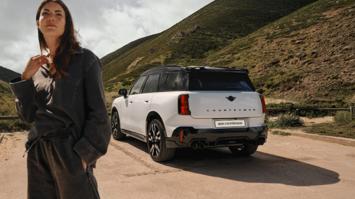 Na sliki sta oseba in bel avtomobil Mini Countryman na cesti v gorskem okolju. Oseba stoji v ospredju na levi strani slike, avtomobil pa je parkiran na cesti v ozadju. Za njima so vidni pobočje hriba, cesta in skalnato naravno okolje.