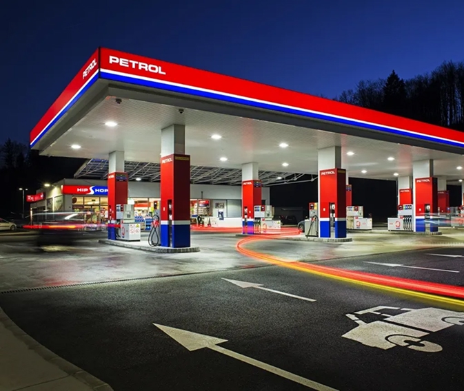Petrolovo prodajno mesto ponoči z osvetljenimi točilnimi mesti, prodajalno in svetlobnimi sledmi vozil.