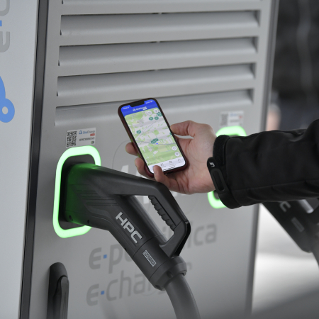 Na sliki je električna polnilnica, ob kateri uporabnik na mobilnem telefonu preverja lokacije polnilnih mest med polnjenjem vozila.
