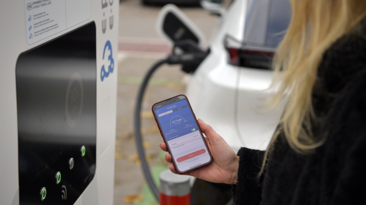 Na sliki uporabnica ob polnjenju električnega vozila uporablja mobilni telefon za identifikacijo in plačilo polnjenja.